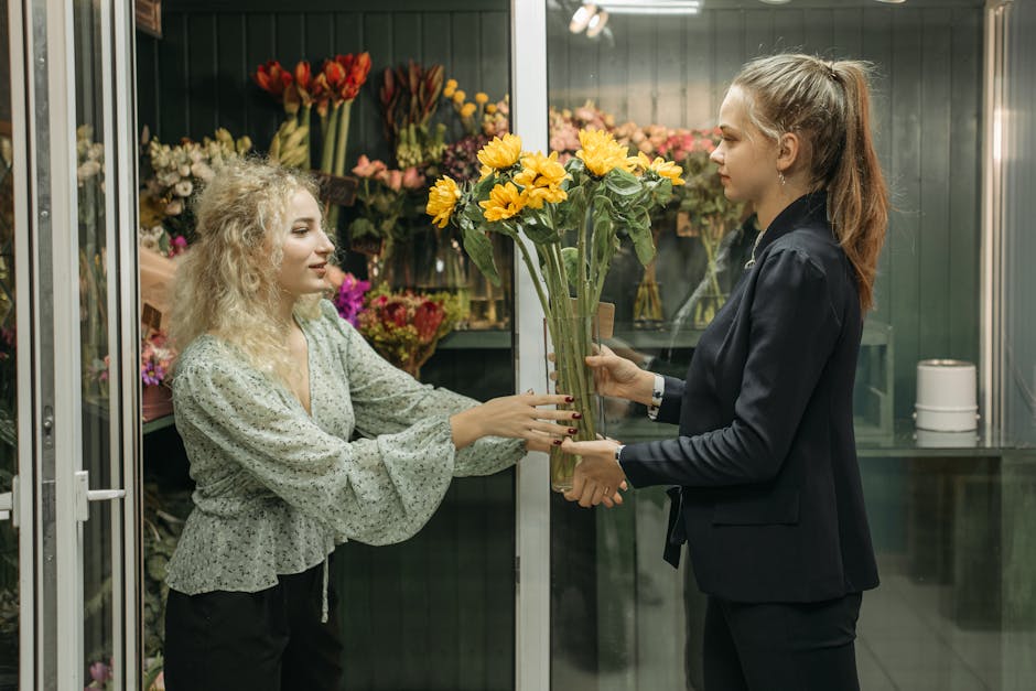 A florist with curly blonde hair, wearing a light green floral blouse, is handing a bouquet of vibrant yellow sunflowers to a customer in a dark blazer with a ponytail inside a flower shop. The bouque