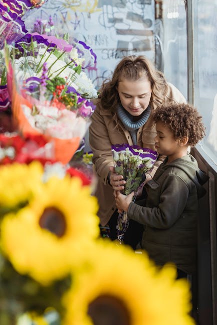A woman and a young girl are inside a flower shop near a large window, engaging in a flower delivery transaction. The woman, with light brown, wavy hair pulled back and wearing a brown coat and a gray