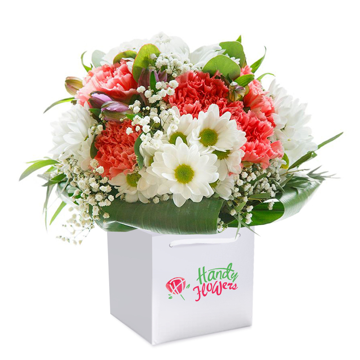 Bathed in soft, natural light and placed upright within a pristine square white gift bag, this vibrant bouquet captures the spirit of Radlett's countryside charm. Fluffy coral and peach carnations unfurl their ruffled petals beside deep red accents, forming a textured tapestry brimming with warmth and affection. Bright, white daisies with playful lime-green centers bring a cheerful contrast, echoing the daisy-studded greenspaces often seen along Theobald Street. Sprigs of baby's breath weave gently through the bouquet, their tiny white clusters adding an airy softness and a touch of nostalgia. On the arrangement's left, several closed buds edged with purple and pink hues evoke the promise of blossoming, as if gathered at the very start of a Radlett spring. Surrounding the blooms is a collar of lush, deep green leaves-broad and gleaming, reminiscent of the protective shade trees that dot local parks. The arrangement comes together in a harmonious, rounded silhouette that showcases skilled craftsmanship and attention to balance. The crisp white bag, designed with sturdy ribbon handles for effortless gifting, rests on a pure white background, isolating the colours and forms for maximum visual impact. Curated by Flowers Radlett, this bouquet is ideal for sending heartfelt wishes, celebrating milestones, or bringing a breath of countryside joy into any Radlett home-its beauty amplified by the knowledge that it was carefully crafted by expert local hands.