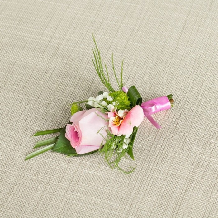Resting diagonally across a beautifully textured, light beige linen, this Radlett-inspired boutonniere seems to glow in gentle afternoon light-a true testament to the artistry of local florists. Dominating the design is a single, immaculate pink rosebud, its petals delicately furled and gradually unfurling outward with subtle gradients from rich rose towards creamy peach. Cradling this centerpiece, a charming peach-tinted alstroemeria unfurls soft petals marked with fine maroon etching and splashes of golden yellow, infusing an extra layer of visual intrigue. Accents of cloud-like baby's breath introduce a delicate, ethereal charm, while textured miniature cushion mums in fresh green punctuate the palette with vibrancy, recalling the woodlands ringing Radlett. The foliage is thoughtfully composed: upright ferny greens and deep-hued folded leaves create a dynamic, layered backdrop, grounding the design and adding organic movement. The stems are elegantly bound by a gleaming fuchsia satin ribbon, tied into a petite bow at the base, which sparkles gently against the understated fabric-lending a festive flourish that feels perfect for a wedding or formal Radlett gathering. The overall arrangement offers not only elegant styling but also evokes the gentle romance and celebration characteristic of local milestone occasions. Each element harmonizes, reflecting an expert hand and an eye attuned to the area's natural beauty, making this boutonniere a fitting accessory for anyone wishing to bring a touch of Radlett's charm to their special day.