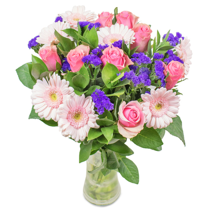 A radiant, expertly arranged bouquet from Flowers Radlett sits elegantly in a clear glass vase, set against a crisp, white studio background. The focal point is a luxurious cluster of seven to nine soft and medium pink roses, petals gently unfurling or just beginning to reveal their classic spiral shape-each one reminiscent of cherished moments in Radlett's welcoming homes. Among these, five to seven pale pink gerbera daisies fan out with large, circular heads and delicate, radiating petals, their lighter centres exuding gentle purity. Vivid purple statice or limonium-clustered in between-introduce a lively textural contrast, adding visual depth and a touch of the unexpected, recalling the wildflowers that grace Radlett's country lanes. Lush, deep green leaves spill around the base, providing vitality and grounding, their freshness suggesting the morning air drifting in from nearby Battlers Green Farm. The transparent vase, displaying pristine stems and clear water, enhances the bouquet's sense of natural abundance and recent harvest. Arranged for occasions such as birthdays, anniversaries, Mother's Day, or to add an elegant flourish to a Radlett office or home, this composition conveys celebration, affection, and the enduring beauty of expertly selected fresh flowers.